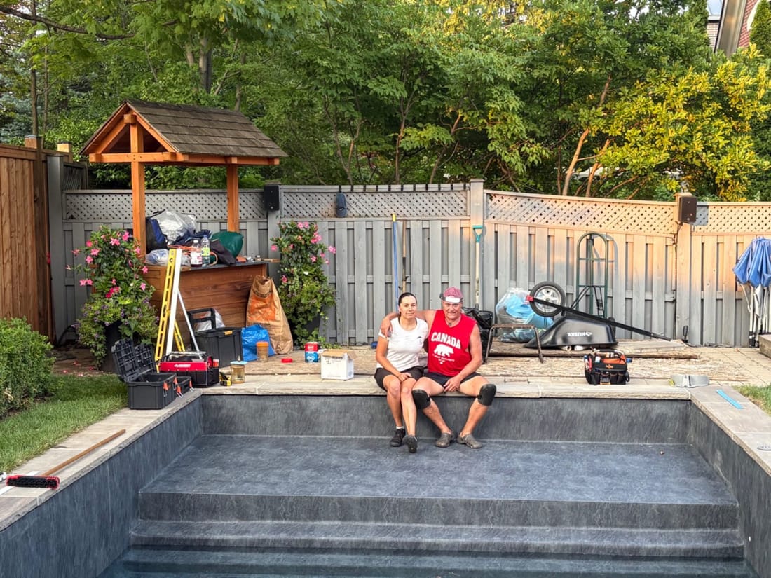 Two people sitting on the edge of a backyard pool lined in grey stone-pattern membrane