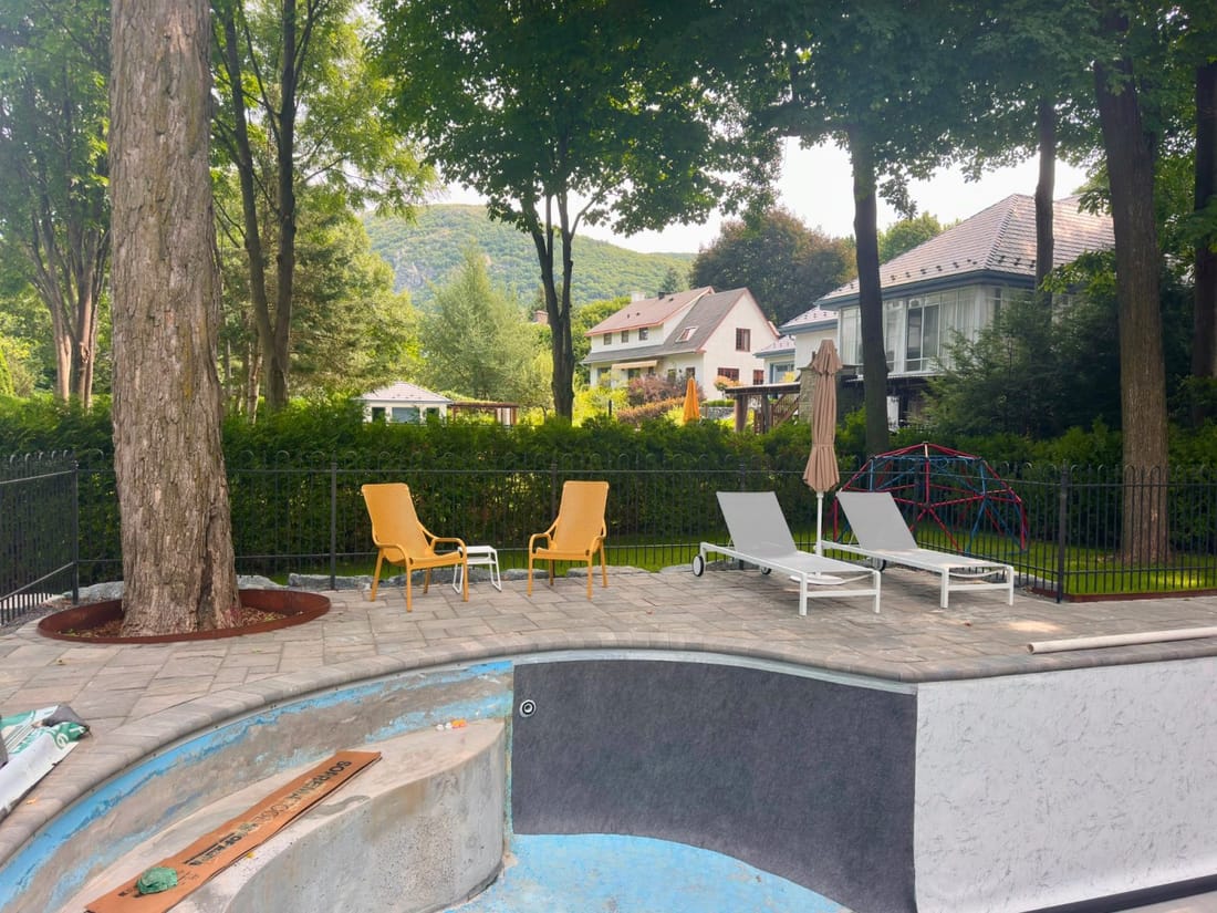 The curved edge of a backyard pool during relining, loungers on the stone deck and trees behind