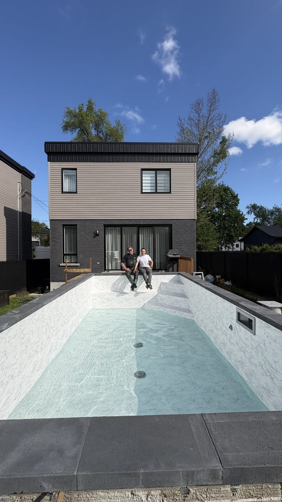 A narrow backyard pool lined in white marble-pattern membrane with a little water on the floor, a two-storey house behind it and two people sitting on the coping at the far end