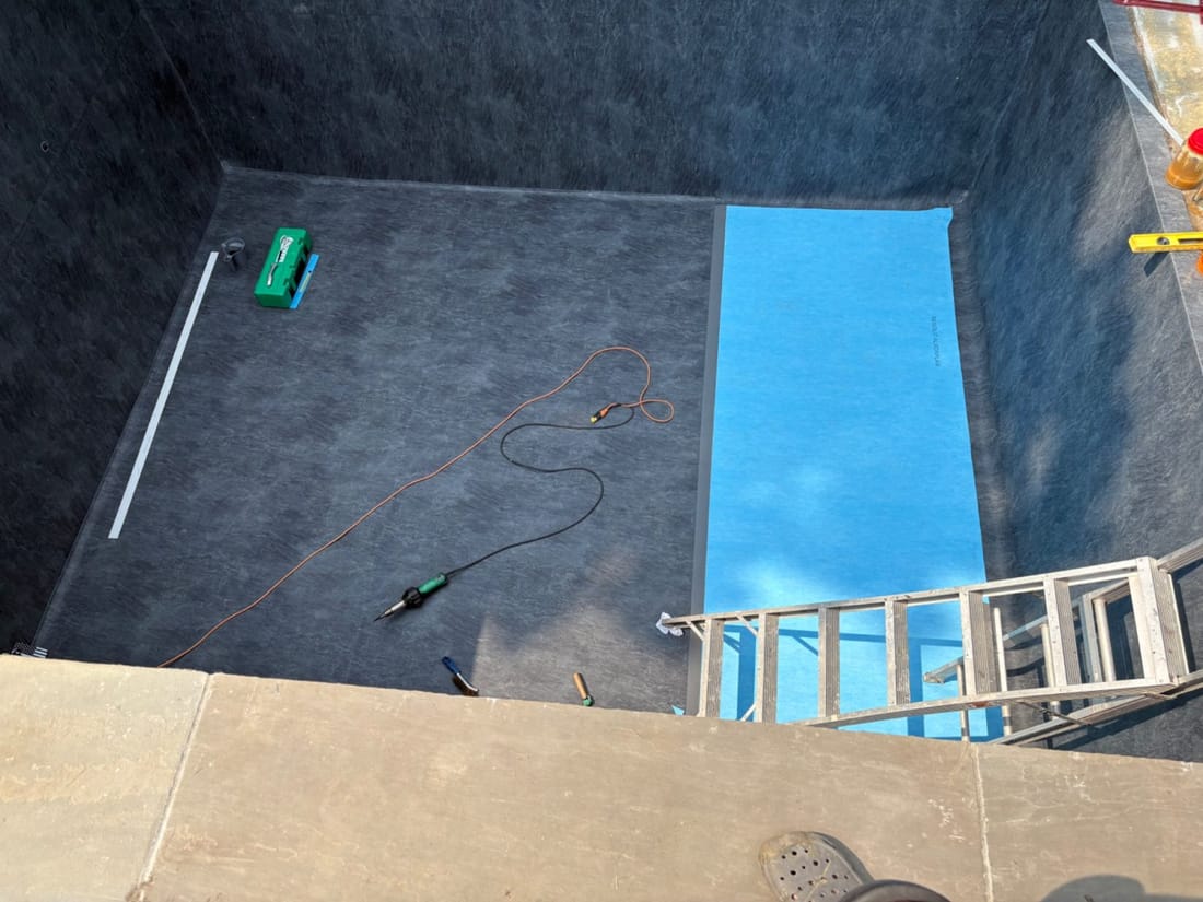 Inside an empty pool: grey geotextile underlay covers the floor and walls, one blue membrane sheet is laid along the right side, and a hot-air welder lies on the floor