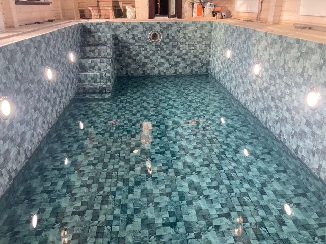 Indoor pool lined in grey-blue mosaic-pattern reinforced PVC membrane, the underwater lights switched on